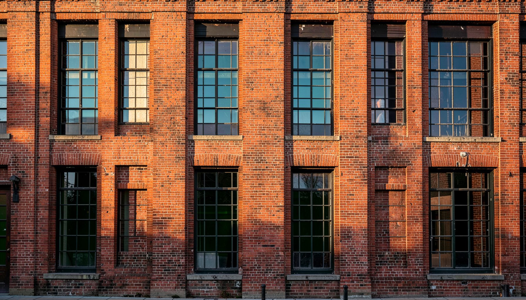 A historic brick warehouse facade in Baltimore with large multi-pane windows, warm terracotta and deep brown tones, early morning light casting long shadows across the weathered masonry